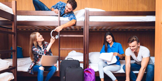 Four young individuals, each on a bunk bed, engaged in various activities – laptop use, music listening, reading, and an ambiguous activity. Suitcases and backpacks suggest a shared living space, possibly a hostel.